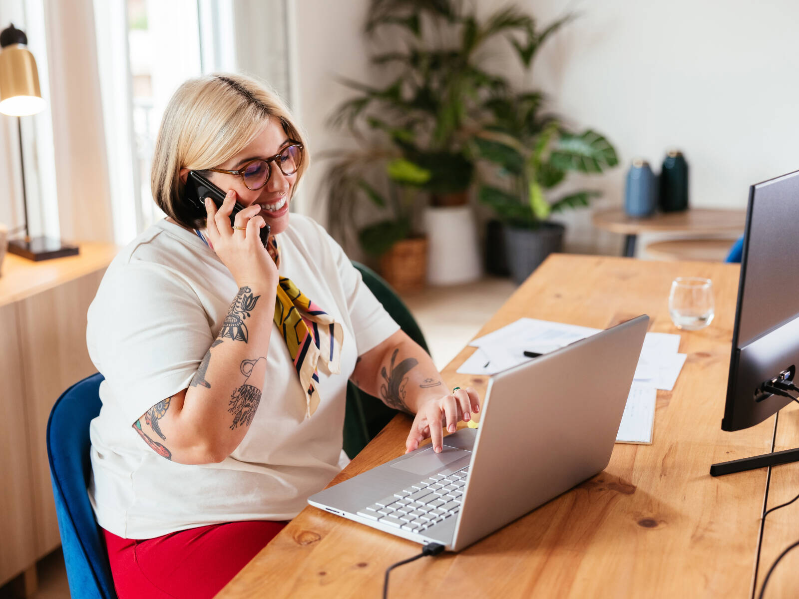 Woman with blonde hair and tattoos talking on the phone while working on a laptop at a wooden desk.