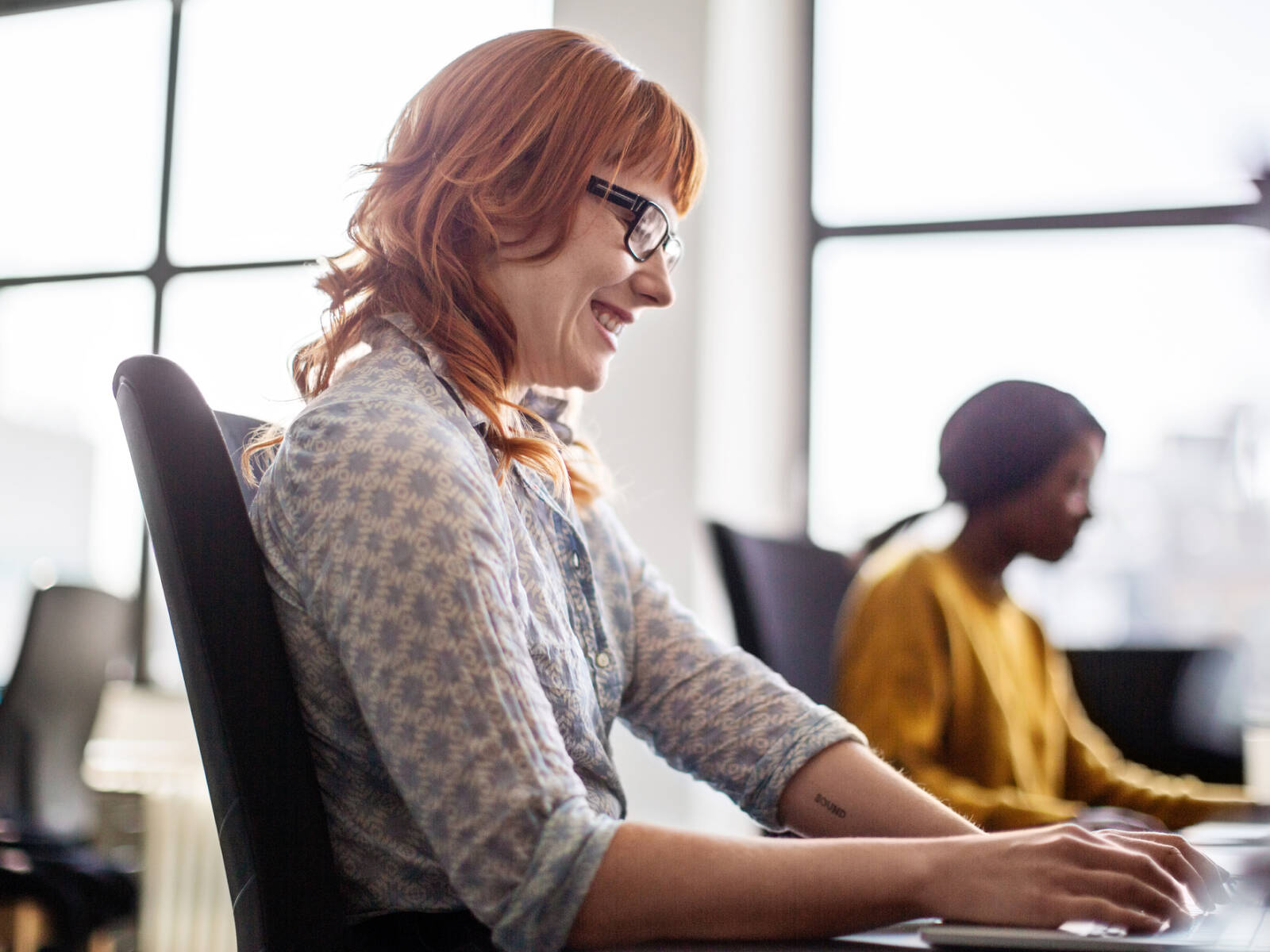 Smiling woman working on a laptop in a bright office, with another colleague working in the background.