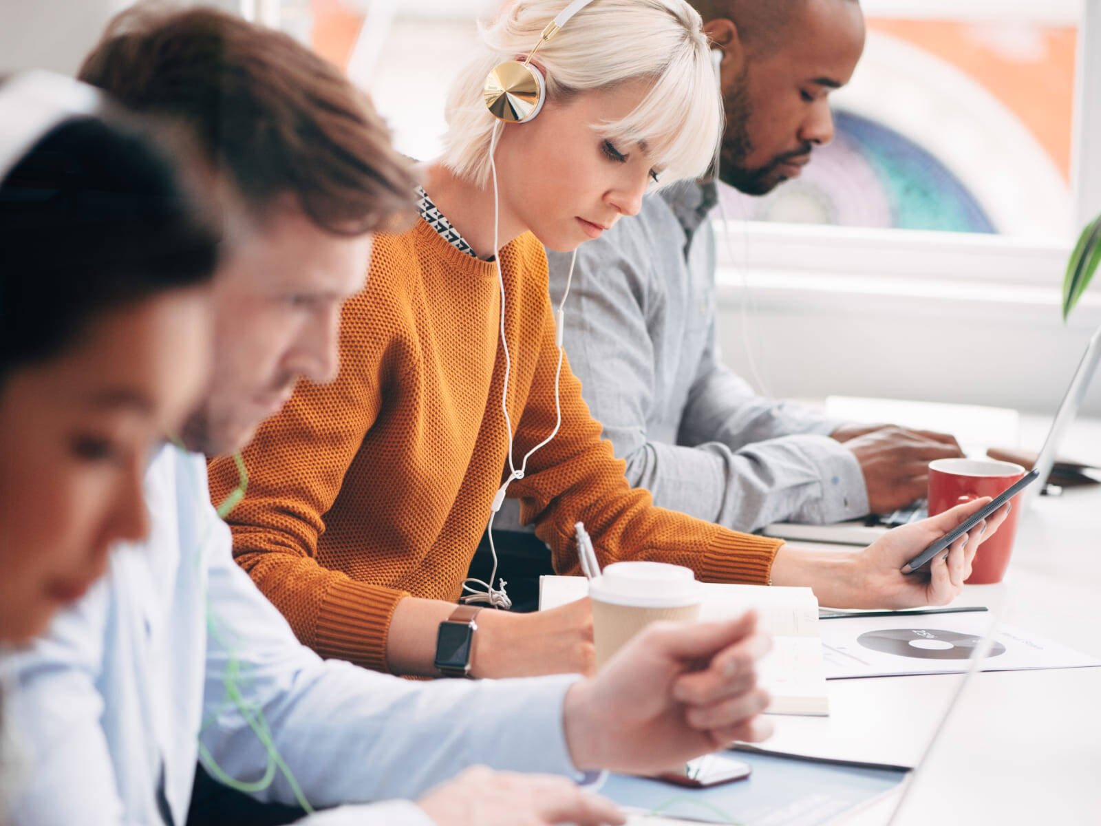 Group of professionals wearing headphones and working on laptops and tablets in a bright collaborative workspace.
